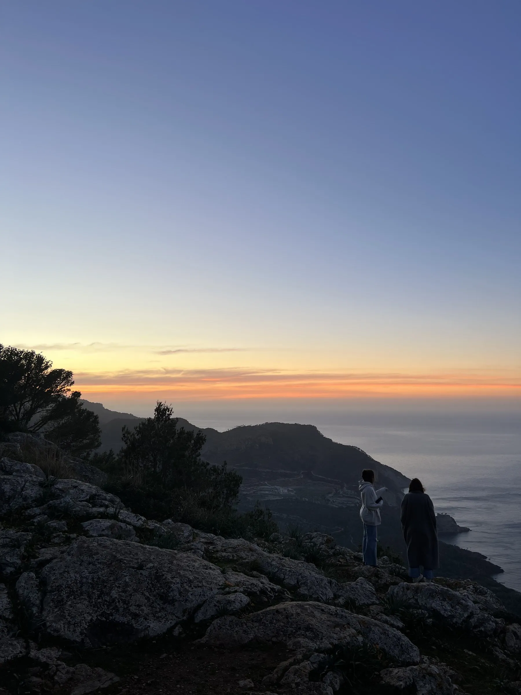 Zwei Personen stehen auf den Felsen und blicken aufs Meer von Banyalbufar, während das Licht schwindet.