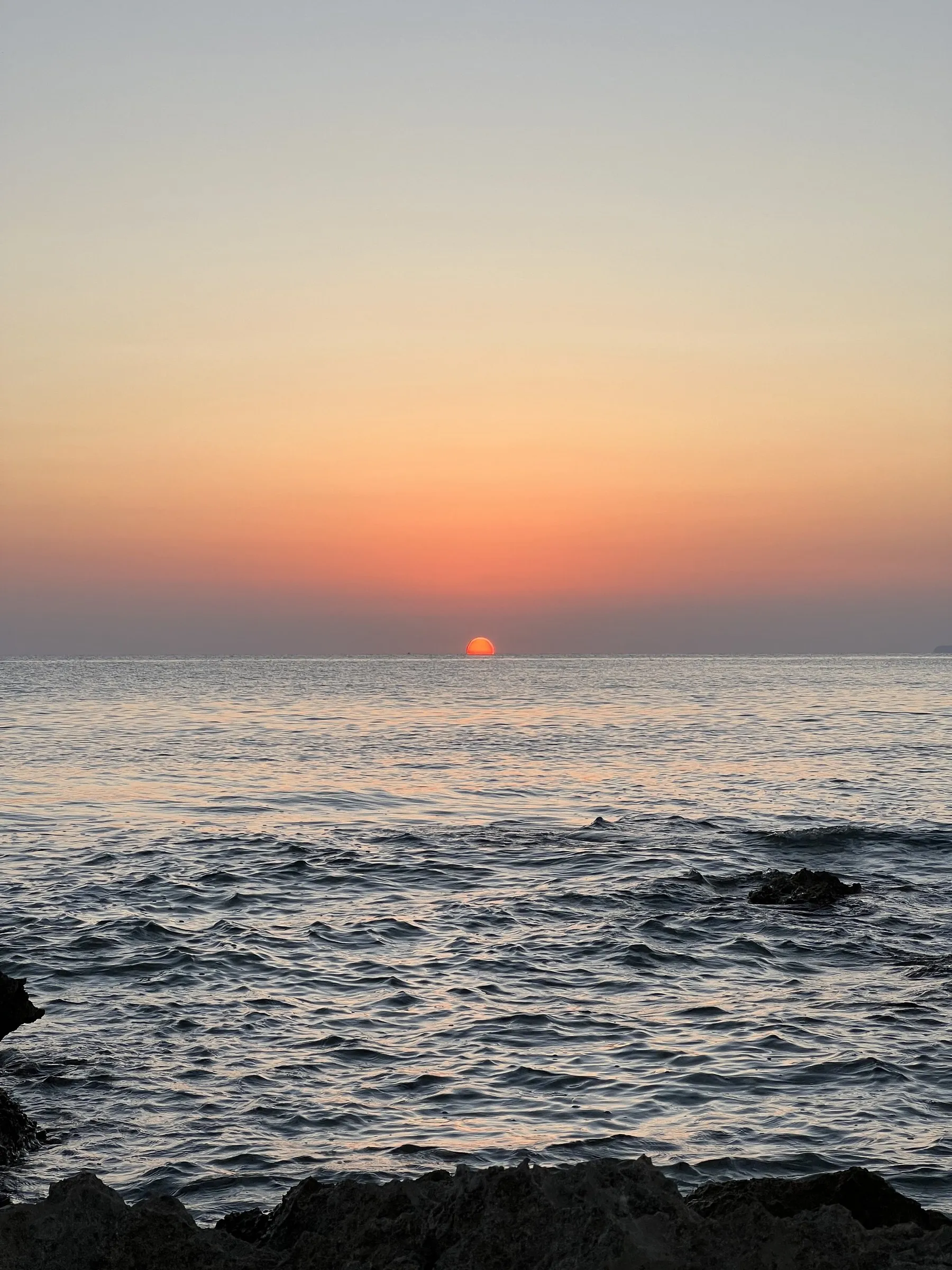 De zon raakt de horizon boven de open zee bij Cap de ses Salines, met lage rotsen op de voorgrond.